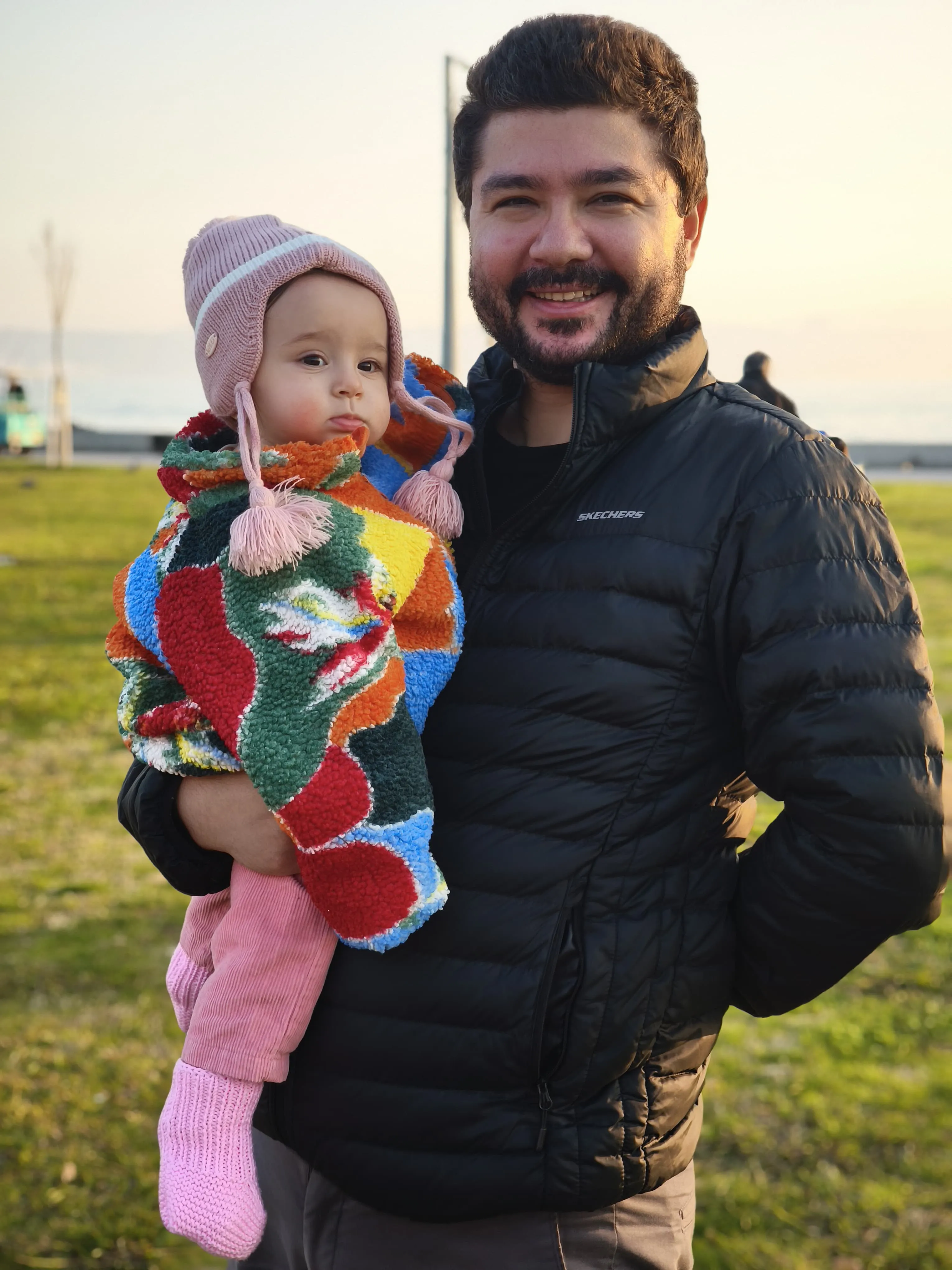 Father holding baby girl outdoors, wearing a colorful fleece blanket and pink hat.