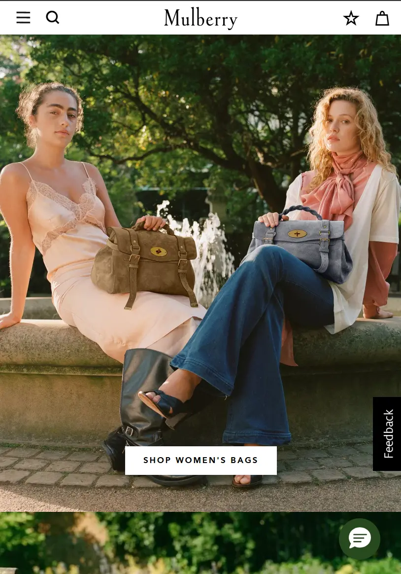 Mulberry website homepage showing two women seated on a stone fountain edge in a sunlit park, one holding a brown suede Alexa satchel and the other a blue-grey one, above a "Shop Women's Bags" button.